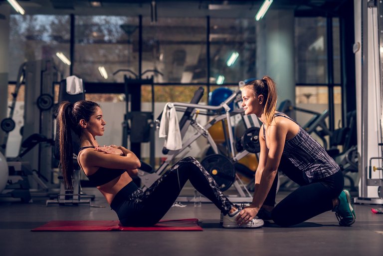 Side view of young motivated attractive healthy sporty active shape girl doing abs exercises and warming on the red mat while her young personal female trainer holding legs in the gym.